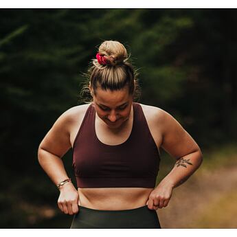Žena ve sportovní podprsence si upravuje kraťasy bjež, s pohledem dolů, stojí v přírodě..